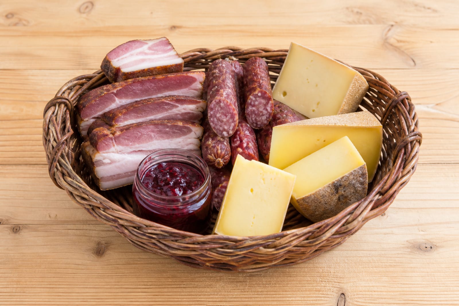 Weidenkorb mit Speck, Würsteln, Käse und Gläsern, in Stroh gebettet