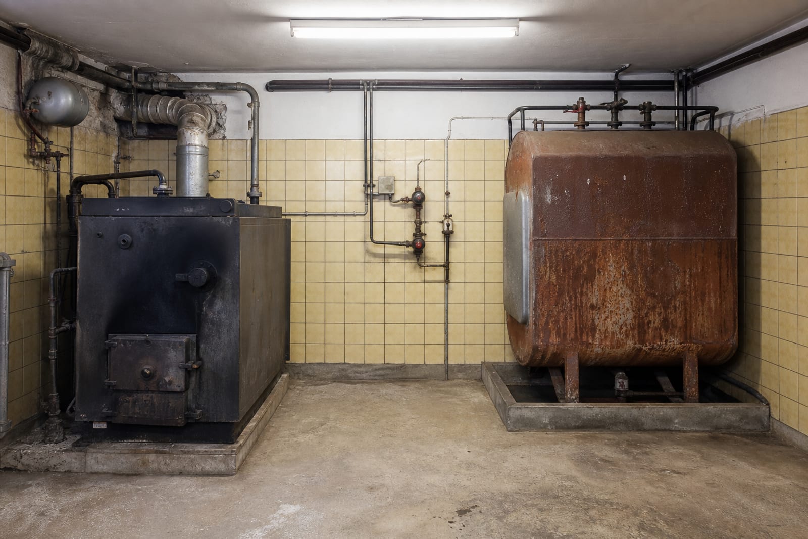 Alter Heizraum mit rußgeschwärztem Ölkessel, verrostetem Stahltank und gelblichen Wandfliesen