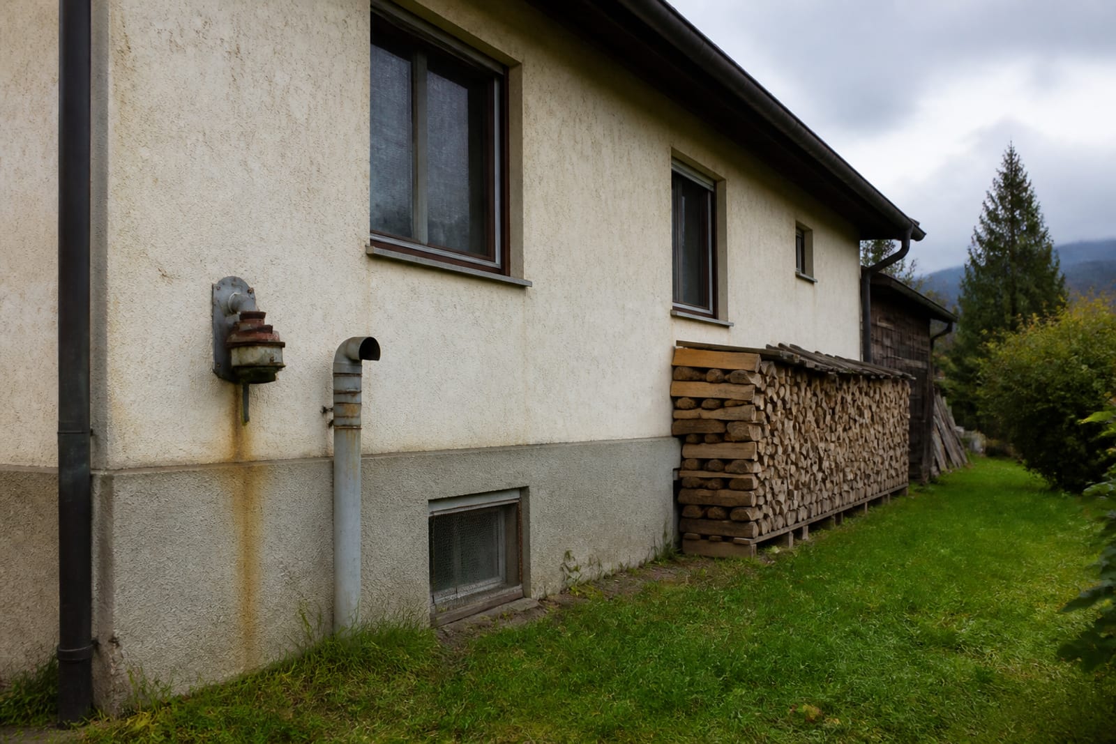 Hausfassade ohne Wärmepumpe: nur ein alter Öltank-Belüftungsstutzen und gestapeltes Brennholz an der Wand